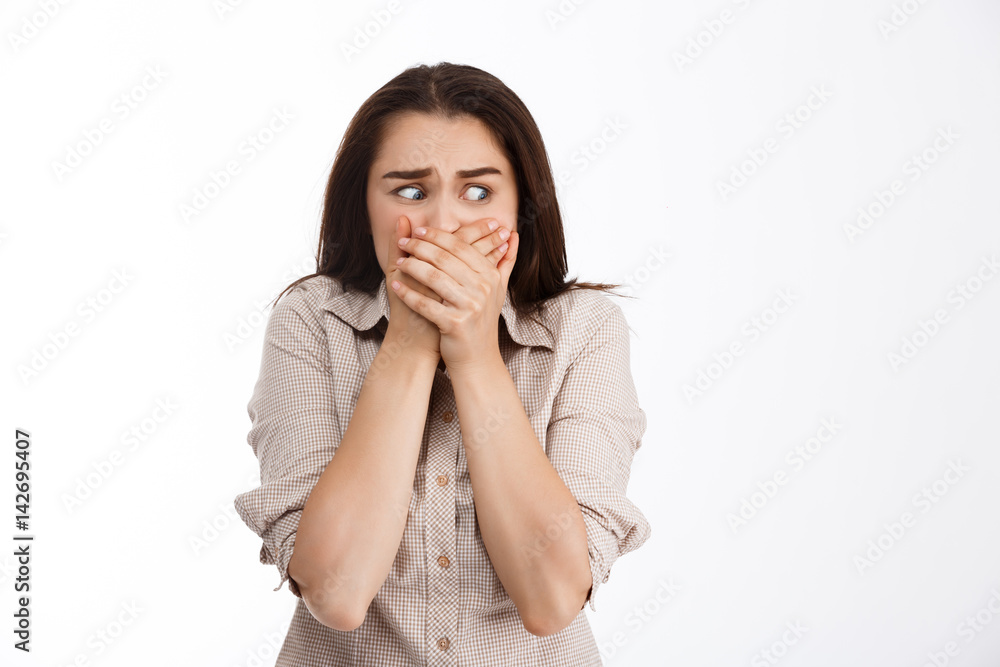 Close up portrait of beautiful scared brunette girl covering her mouth with hands looking away over white background Close up portrait of beautiful scared brunette girl covering her mouth with hands looking away over white background