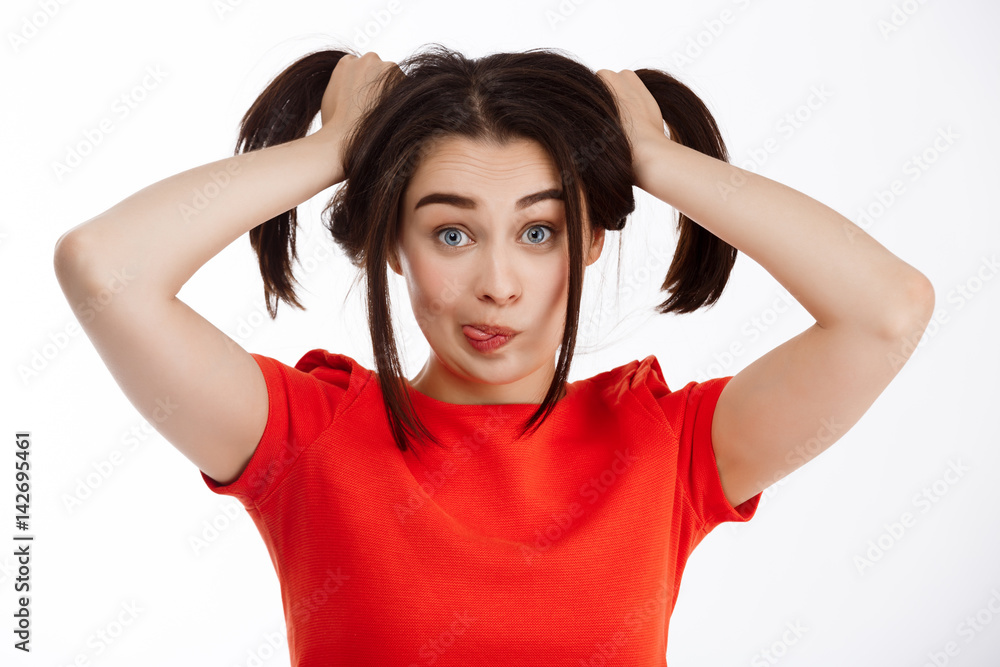 Young beautiful funny crazy brunette girl making pony-tails looking at camera smiling and showing tongue over white background Young beautiful funny crazy brunette girl making pony-tails looking at camera smiling and showing tongue over white background