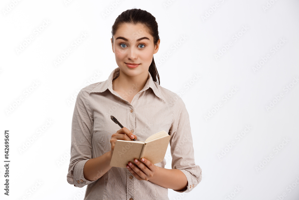 Confident young beautiful businesswoman writing in notebook looking at camera smiling over white background Confident young beautiful businesswoman writing in notebook looking at camera smiling over white background