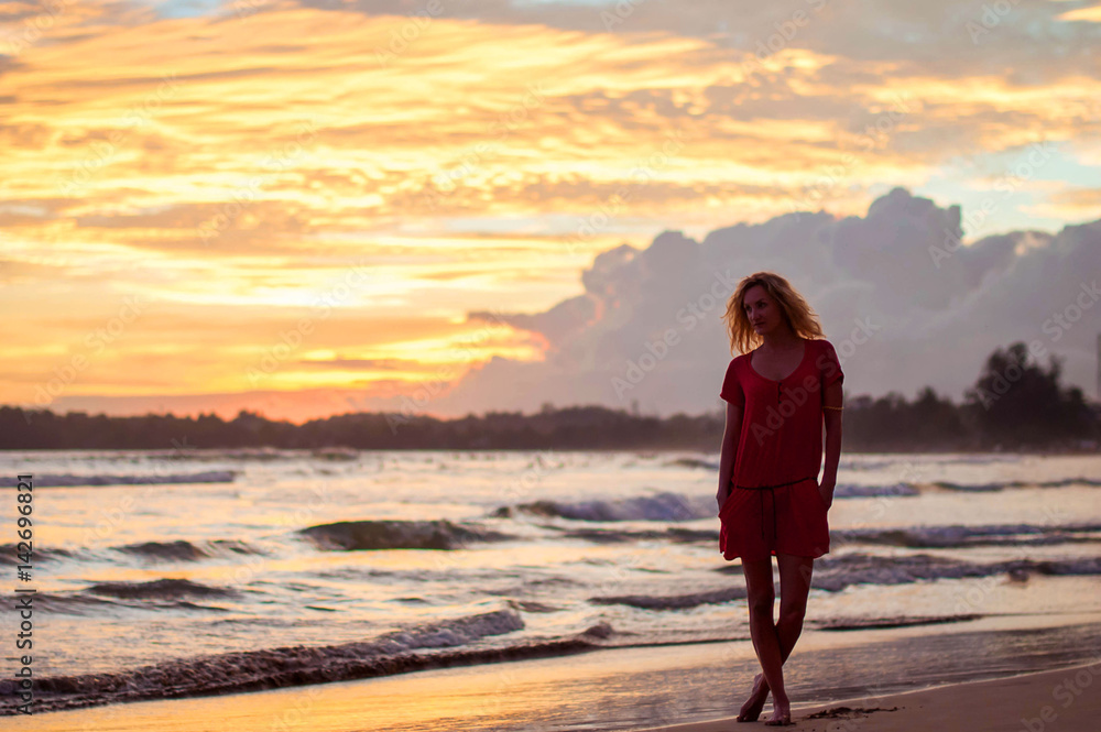 Romantic blond girl in red dress walking on the ocean shore during sunset.