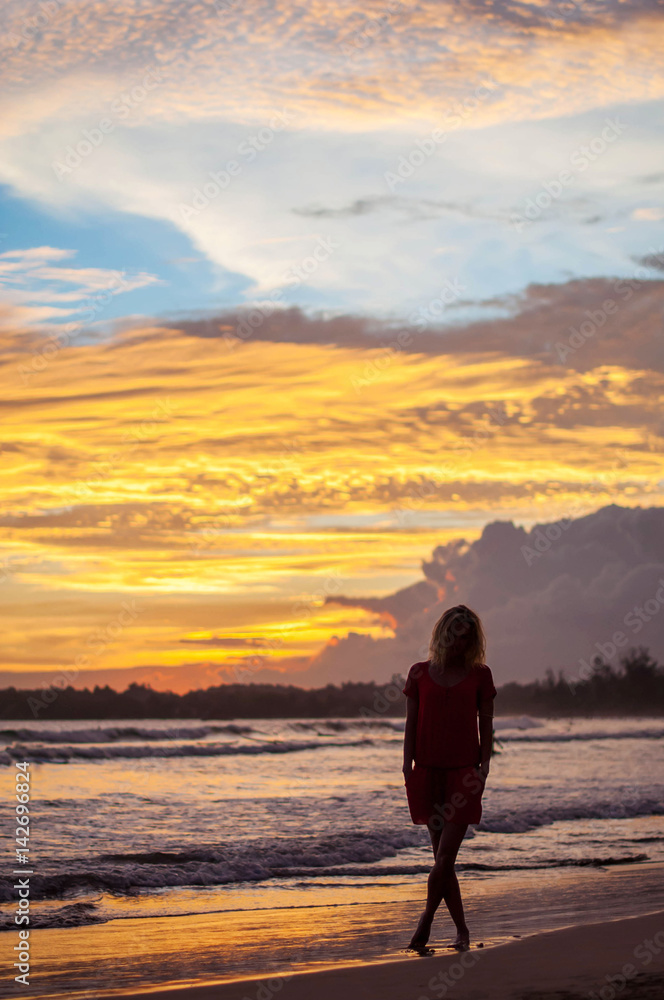Romantic blond girl in red dress walking on the ocean shore during sunset.
