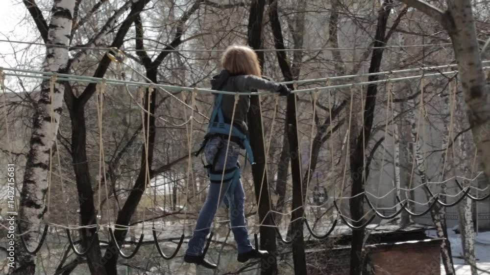 Young female climber walks by pendant rope bridge on high ropes course in extreme park. Rock climbing concept Young female climber walks by pendant rope bridge on high ropes course in extreme park. Rock climbing concept