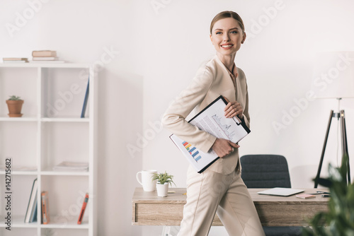 Young smiling businesswoman holding clipboard and looking away in office