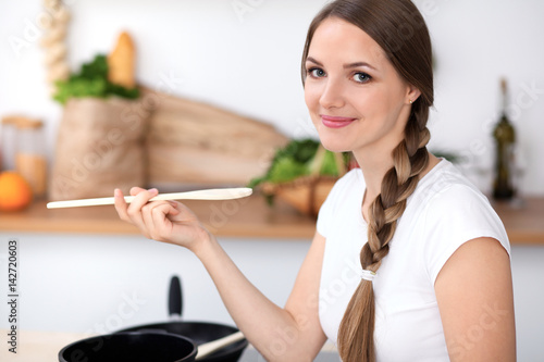 Young woman is cooking in a kitchen. Housewife is tasting the soup by wooden spoon.