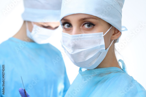 Close up of surgeon woman looking at camera while colleagues performing in background in operation room  isolated. Surgery and medicine concept