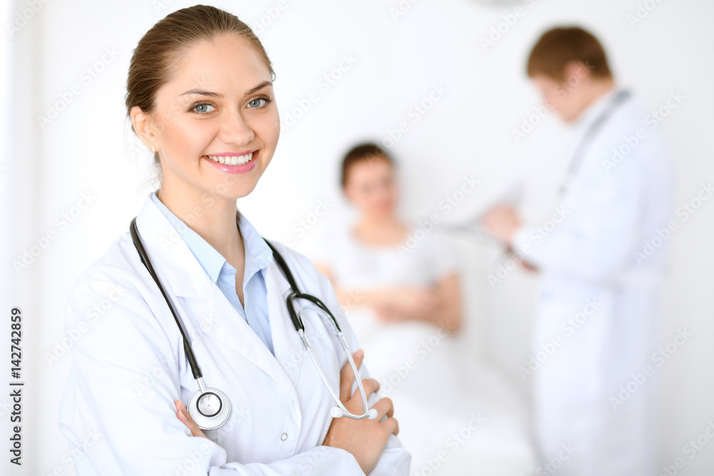 Cheerful smiling female doctor on the background with physician and his patient in the bed. High level and quality of medical service concept. Cheerful smiling female doctor on the background with physician and his patient in the bed. High level and quality of medical service concept.
