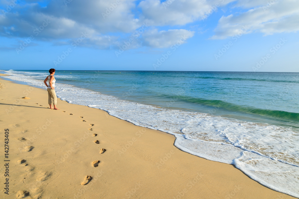 Footprints in sand with young woman tourist standing on beach in Morro Jable, Fuerteventura island, Spain