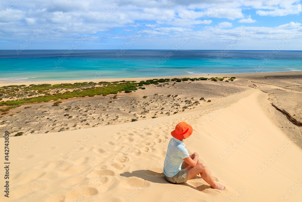 Young woman in orange hat sitting in sand on a dune looking at Sotavento beach on Jandia peninsula, Fuerteventura, Canary Islands, Spain Young woman in orange hat sitting in sand on a dune looking at Sotavento beach on Jandia peninsula, Fuerteventura, Canary Islands, Spain
