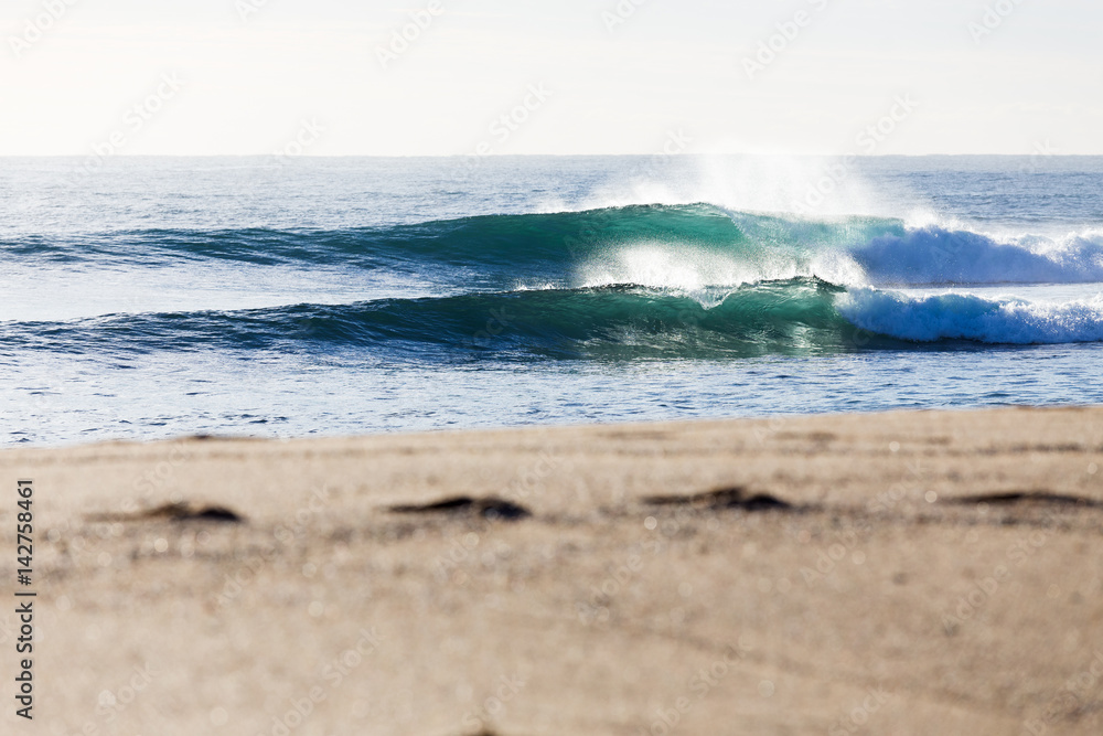 Fresh ocean waves breaking in bright morning light towards a beautiful sandy beach with footprints in the foreground.