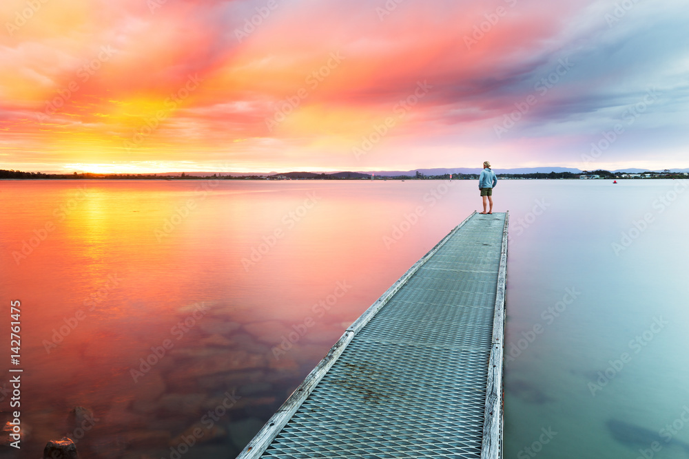 A person watches a beautiful, colourful sunset over the sea from the end of a small jetty. A person watches a beautiful, colourful sunset over the sea from the end of a small jetty.
