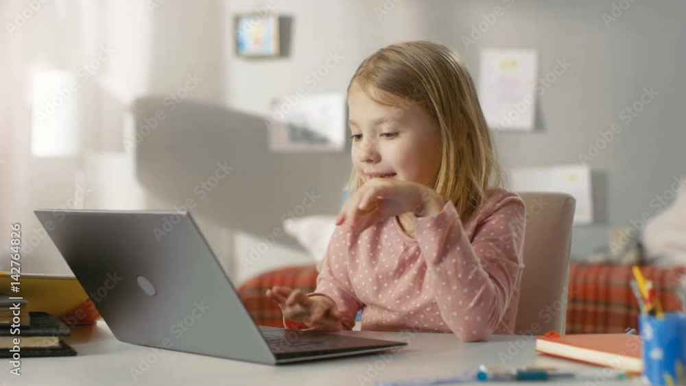 Cute Young Girl Uses Laptop Computer with Interest while Sitting at the Table in Her Light Room. Shot on RED EPIC-W 8K Helium Cinema Camera. Cute Young Girl Uses Laptop Computer with Interest while Sitting at the Table in Her Light Room. Shot on RED EPIC-W 8K Helium Cinema Camera.