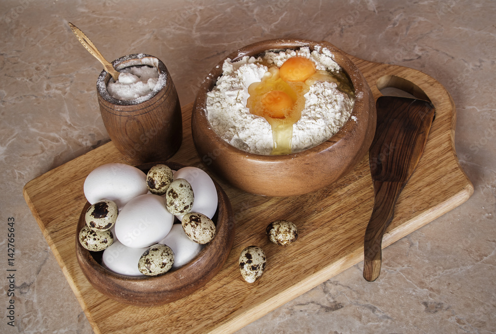 chicken and quail eggs in wooden bowl and bowl of flour and salt on stone surface kitchen table top view