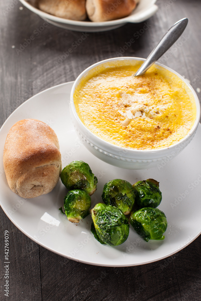 Roasted Butternut Squash Soup with shaved Parmigiano Reggiano Cheese and cinnamon on a dark wooden background served with roasted Brussels sprouts and a honey wheat roll side view Roasted Butternut Squash Soup with shaved Parmigiano Reggiano Cheese and cinnamon on a dark wooden background served with roasted Brussels sprouts and a honey wheat roll side view