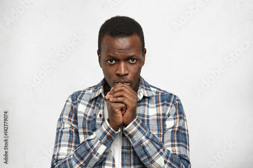 Handsome young dark-skinned man in casual shirt keeping hands in front of him while praying, hoping for best, worrying about poor state of health of his wife, having worried and stressed look