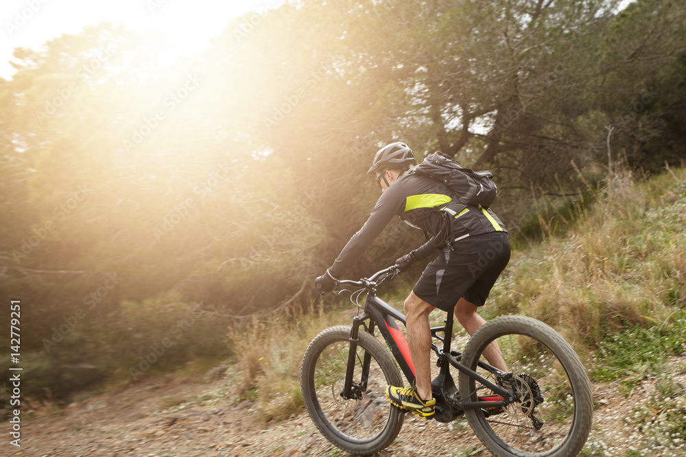 Rear shot of European professional rider in helmet, cycling clothes and eyeglasses riding black electric bike down trail with sun shining ahead of him. People, active healthy lifestyle and extreme