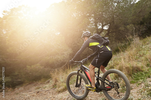 Rear shot of European professional rider in helmet  cycling clothes and eyeglasses riding black electric bike down trail with sun shining ahead of him. People  active healthy lifestyle and extreme