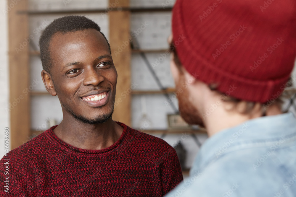 Obraz premium People, interracial friendship and communication concept. Handsome cheerful young Afro-American man dressed in cozy sweater smiling happily rejoicing at that his Caucasian friend got married
