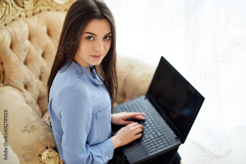 Fotografie brunette in blue shirt is sitting in a vintage armchair by the window with lapto