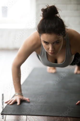 Young resolute woman practicing yoga, doing Push ups or press ups exercise, working out on black mat, close up portrait, loft background. Weight loss, healthy lifestyle concept, vertical photo