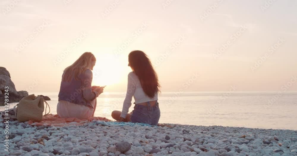 Two Women sitting on beach using smartphone smiling and texting best girl friends with mobile phone browsing social media for inspirational travel photos enjoying a relaxing summer vacation at sunset on beach Two Women sitting on beach using smartphone smiling and texting best girl friends with mobile phone browsing social media for inspirational travel photos enjoying a relaxing summer vacation at sunset on beach