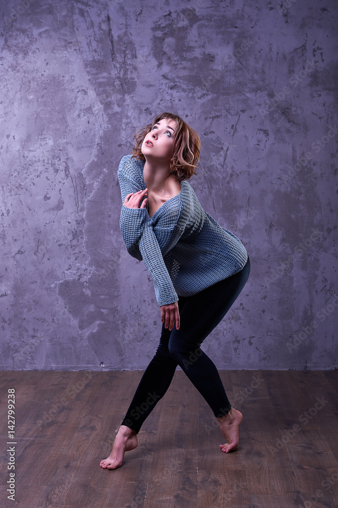 One young beautiful slender dancer girl with short brown curly hair in a grey knitted sweater and black velvet leggings, amazing posing in the dance, barefoot in the studio fashion photography.