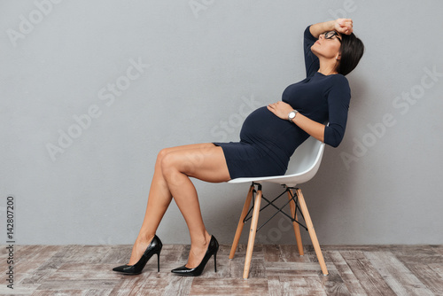 Bored pregnant business woman sitting over grey background.