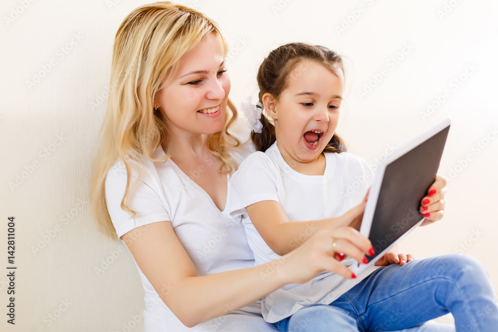 Beautiful young woman and her charming little daughter are using a digital tablet and smiling while lying in bed at home Beautiful young woman and her charming little daughter are using a digital tablet and smiling while lying in bed at home