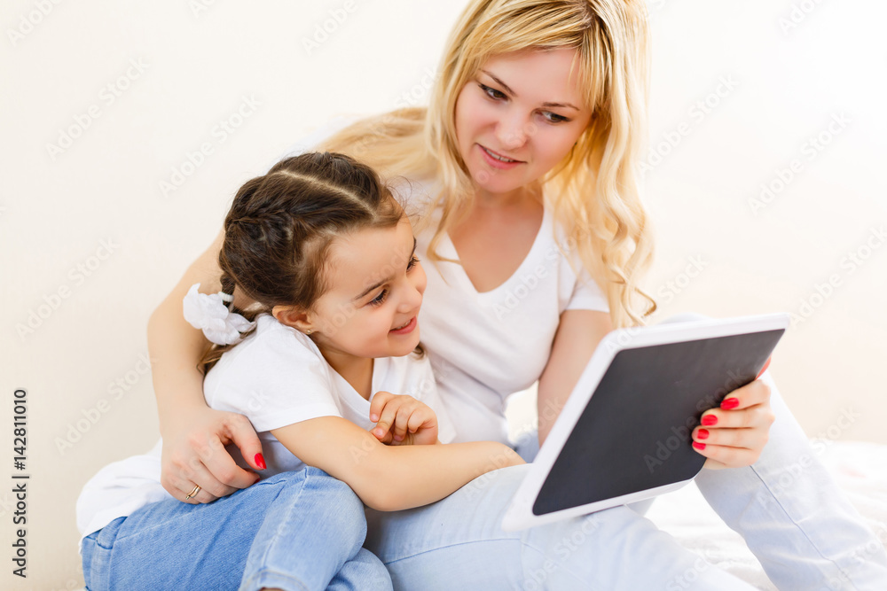 Beautiful young woman and her charming little daughter are using a digital tablet and smiling while lying in bed at home Beautiful young woman and her charming little daughter are using a digital tablet and smiling while lying in bed at home