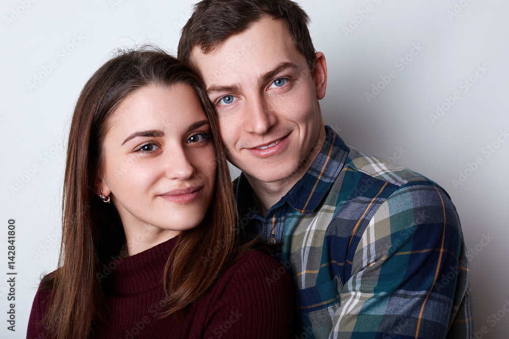 A handsome guy with attractive blue eyes in checked shirt embracing his young pretty girlfriend with brown eyes and nice features. A loving couple standing together hugging each other. A handsome guy with attractive blue eyes in checked shirt embracing his young pretty girlfriend with brown eyes and nice features. A loving couple standing together hugging each other.