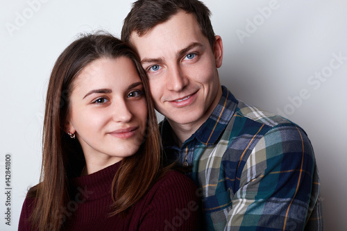 A handsome guy with attractive blue eyes in checked shirt embracing his young pretty girlfriend with brown eyes and nice features. A loving couple standing together hugging each other.