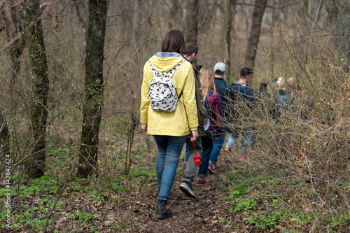 Group of friends walking with backpacks in spring forest from back. Backpackers hiking in the woods. Adventure, travel, tourism, active rest, hike and people friendship concept.