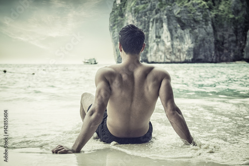 Full body back shot of a handsome young man sitting on a beach in Phuket Island  Thailand  shirtless wearing boxer shorts  showing muscular fit body