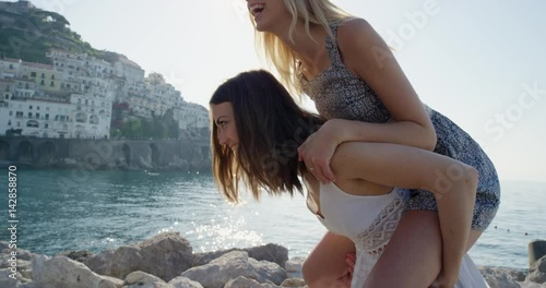 Two girl friends giving piggyback on vacation Tourist woman laughing and walking on beach promenade on summer holiday European travel adventure Amalfi Coast Italy