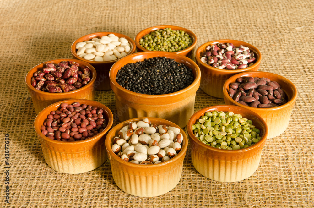 9 pottery containers with various types of pulse and legume seeds on a burlap background in late afternoon light.