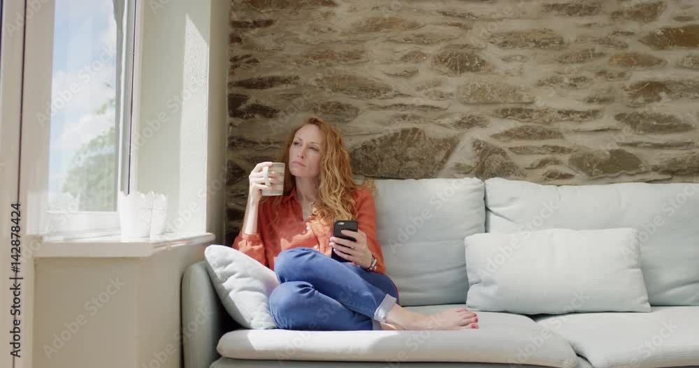 Beautiful redhead woman using smartphone technology at home drinking coffee touching screen browsing social media for authentic creative content Beautiful redhead woman using smartphone technology at home drinking coffee touching screen browsing social media for authentic creative content