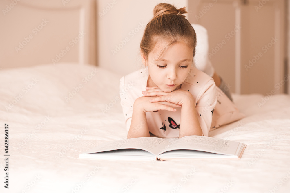 Cute baby girl lying in bed reading book in room. Wearing pajamas at home. Childhood. Cute baby girl lying in bed reading book in room. Wearing pajamas at home. Childhood.