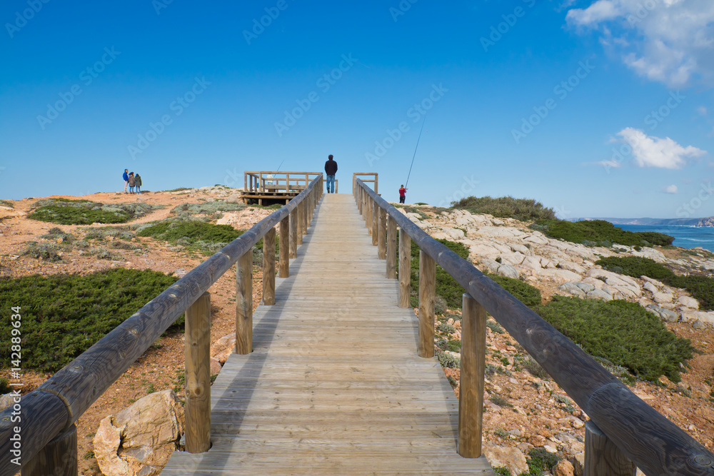 Obraz premium tourists visiting, sightseeing and going up to the point of view on wooden pathway by the atlantic ocean in colorful scenery blue sky, algarve, portugal