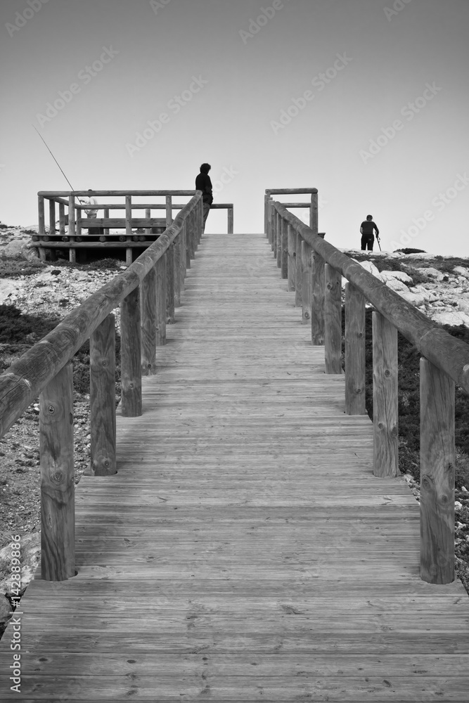 visiting, sightseeing and going up to the point of view on wooden pathway by the atlantic ocean in black and white, algarve, portugal
