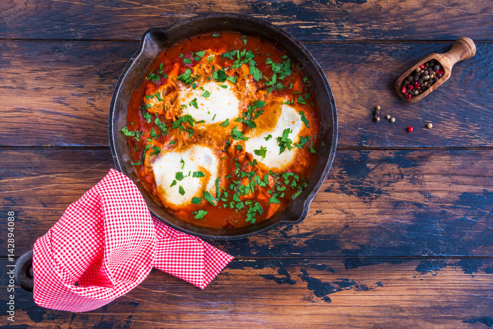 Traditional Arabic dish shakshuka in a black cast iron pan and pepper on the wooden brown background, top view.