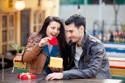 photo of cute couple sitting on the bench and looking at the heart shaped toy on the wonderful cafe background