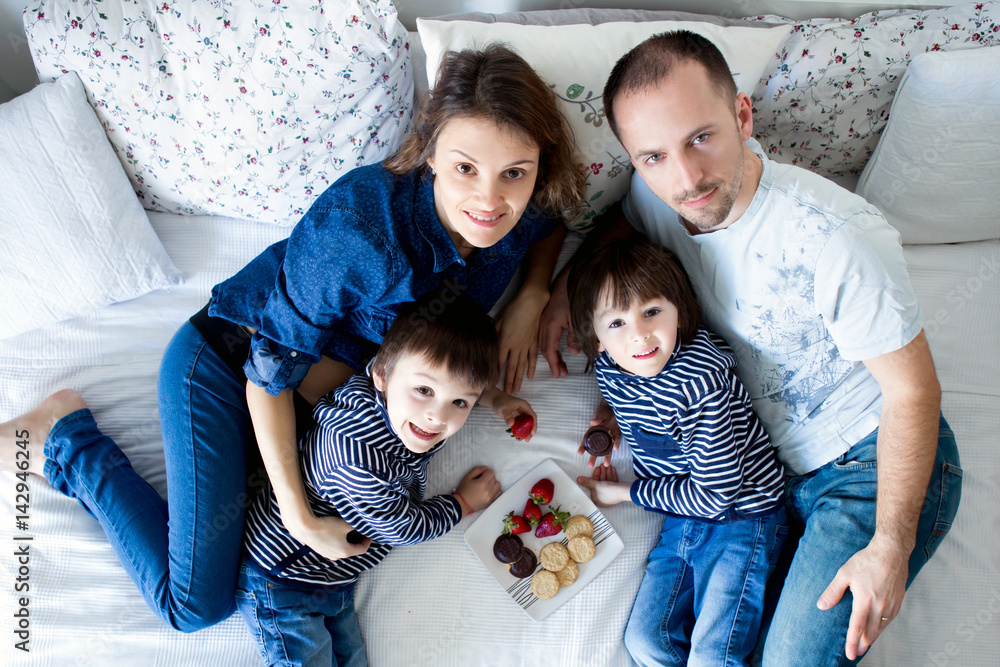 Beautiful family of four, lying on the bed, eating strawberries and cookies