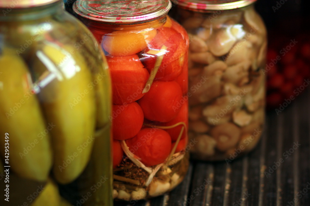 Row of jars with pickled tomatoes, cucmbers and mushrooms standing on black wooden floor Row of jars with pickled tomatoes, cucmbers and mushrooms standing on black wooden floor