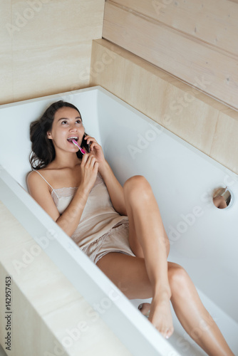 Woman talking on cell phone and brushing teeth in bathtub