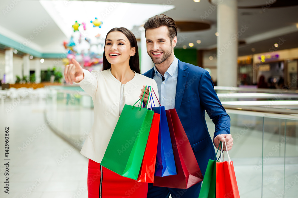 It's shopping time with sales and fun. Cute cheerful successful happy lovely couple in jacket holding colored shopping bags, pointing on shop and laughing in mall at holiday It's shopping time with sales and fun. Cute cheerful successful happy lovely couple in jacket holding colored shopping bags, pointing on shop and laughing in mall at holiday