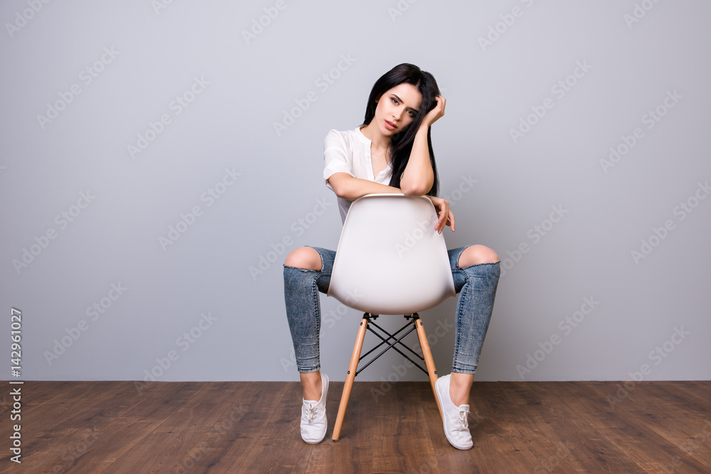 Fototapeta premium Portrait of beautiful sexual young woman posing and touching black hair while sitting on white modern chair