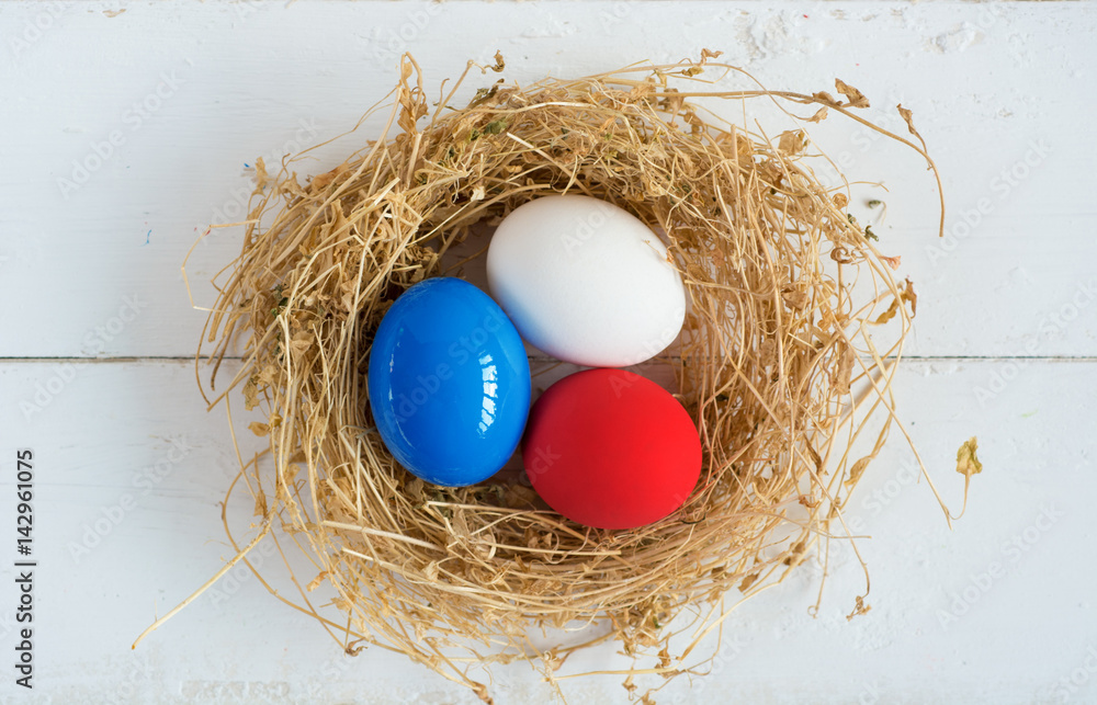 Fototapeta premium Red, blue, white eggs ( As flag of some countries ) in nest on rustic wooden background, selective focus image. Happy Easter Card - space for text. 