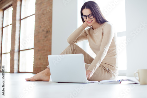 Portrait of beautiful young smiling woman sitting on the floor at home and typing on the laptop