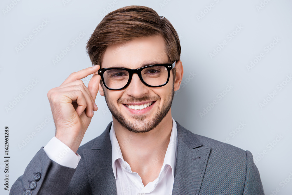 Portrait of successful handsome young man in formal wear with beaming smile and bristle holding glasses while standing on gray background Portrait of successful handsome young man in formal wear with beaming smile and bristle holding glasses while standing on gray background