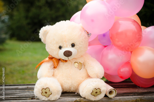 Beige bear on the bench, around multi-colored balloons. Gift for birthday