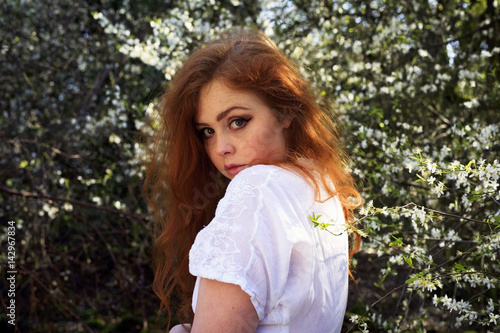 Beautiful portrait of a young woman with long red hair, freckles and sultry pensive looks, set against a woodland background with white blossom